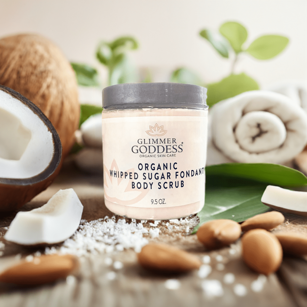Jar of Glimmer Goddess Organic Whipped Sugar Fondant Exfoliating Body Scrub surrounded by coconut and almond, highlighting natural ingredients for effective skincare.