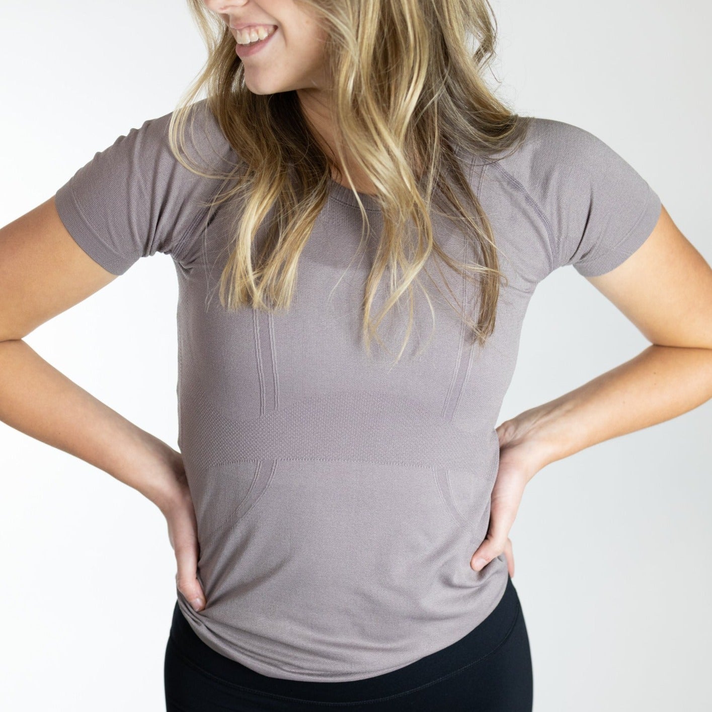 Smiling woman wearing the Trainer Lightweight Short Sleeve Shirt by DYM Athletics in a light brown shade, demonstrating its comfortable and versatile design.