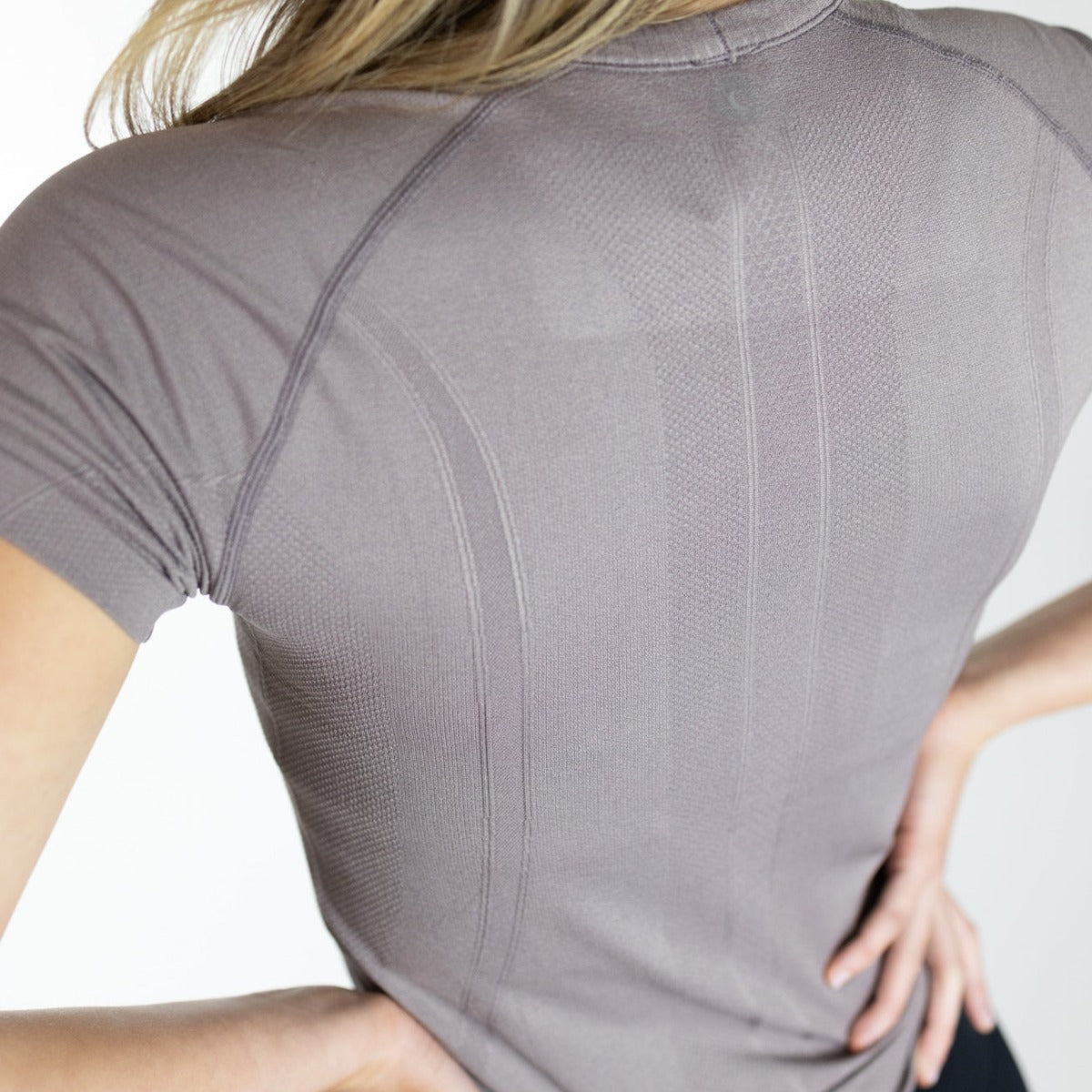 Back view of a woman in a light brown Trainer Lightweight Short Sleeve Shirt by DYM Athletics, emphasizing its detailed seam construction and breathable fabric.