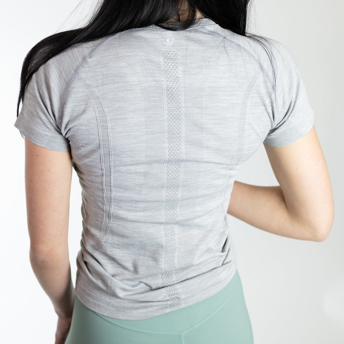 Back view of a woman wearing the Trainer Lightweight Short Sleeve Shirt by DYM Athletics, showcasing its breathable mesh design and flattering seam details.