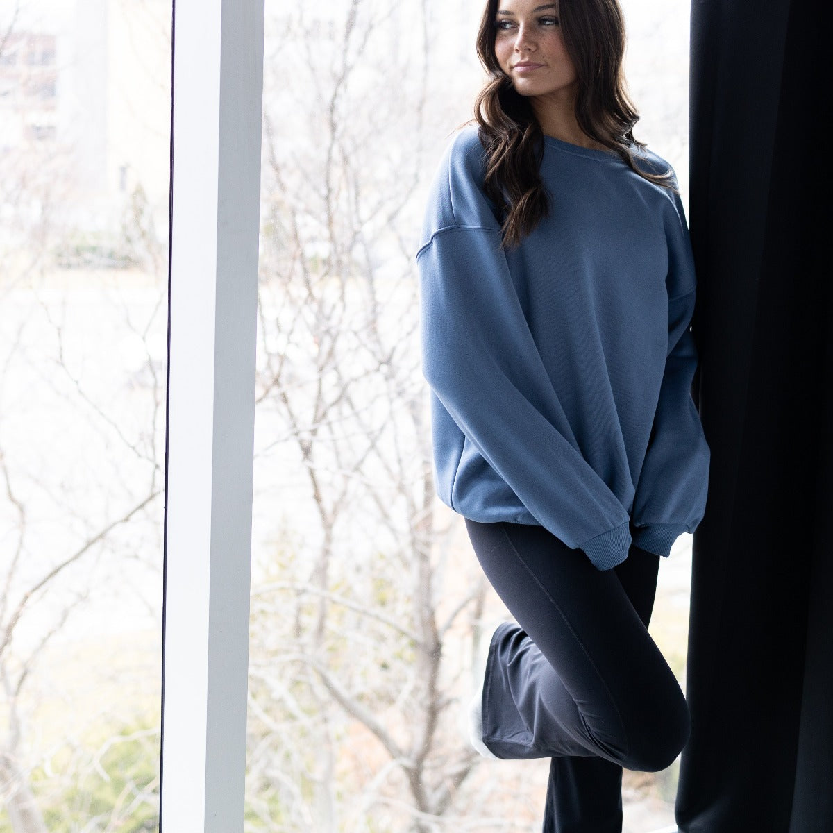Model wearing AIRE Ultra-Soft Flare High Waist Pants by DYM Athletics, showcasing the soft fabric and no front seam design, posed next to a window in a relaxed setting.