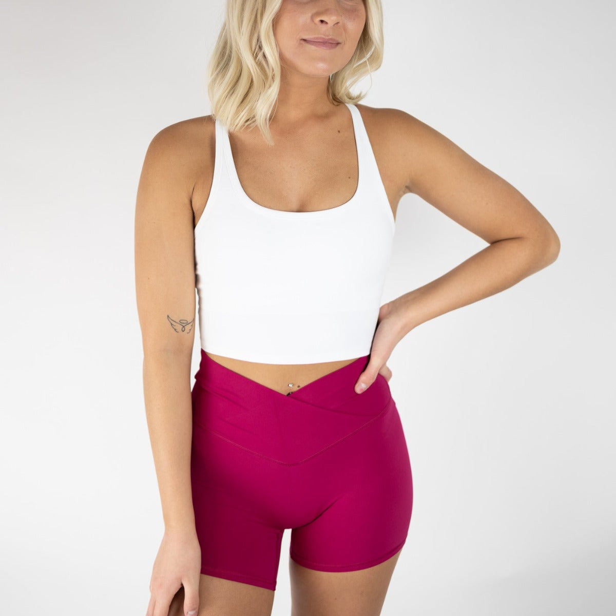 Woman posing confidently in DYM Athletics Flex & Flow 4" V-Waist Shorts in vibrant pink, paired with a white crop top, showcasing comfort and style.