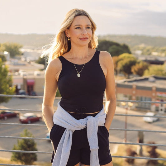 Woman wearing the DYM Athletics Bliss High Neck Bra with Scoop Back in black, styled with a sweater around her waist, enjoying a sunny outdoor setting.