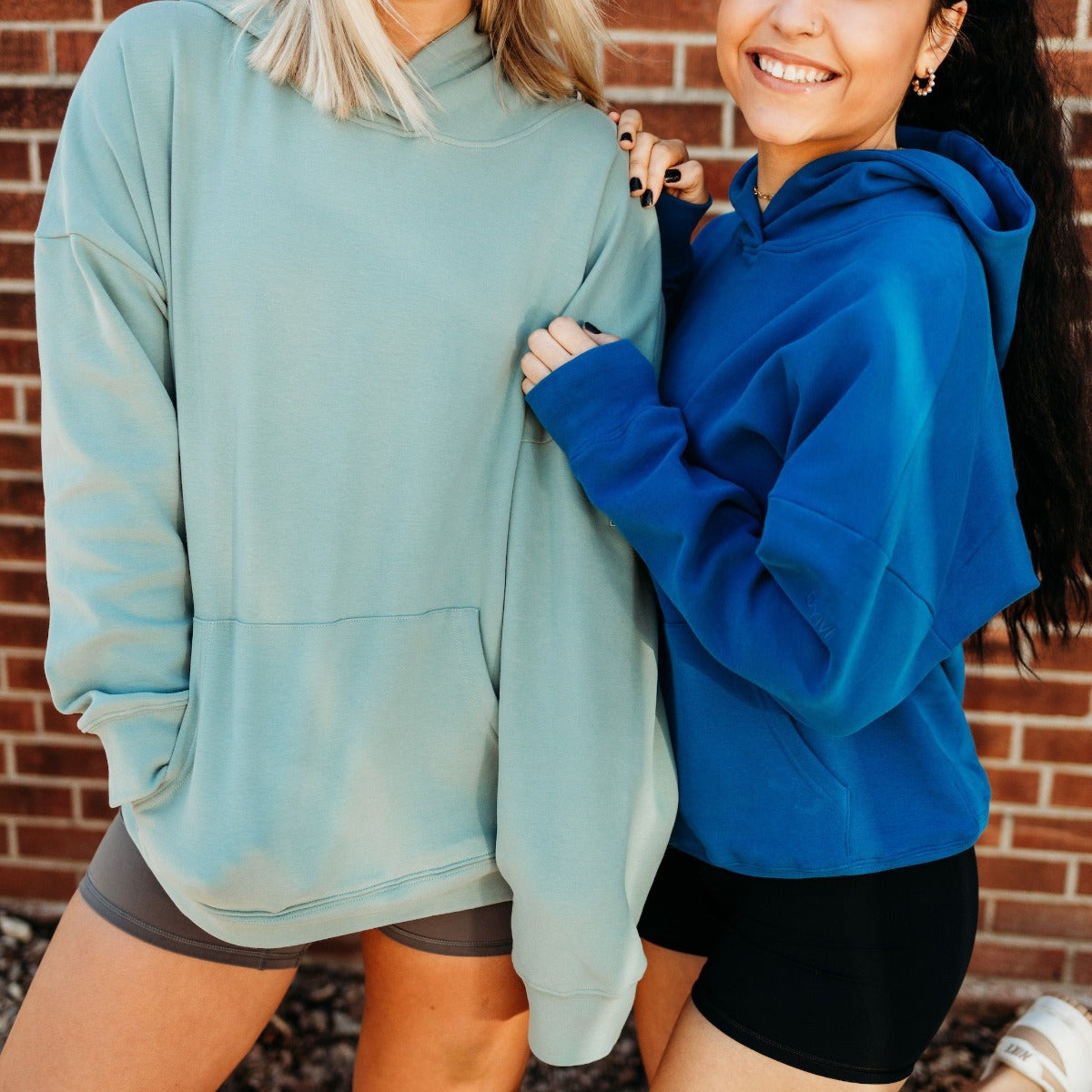 Two models in diverse color options of the Cozy Up Boyfriend Hoodie from DYM Athletics, showcasing their oversized fit and comfort in a friendly pose.