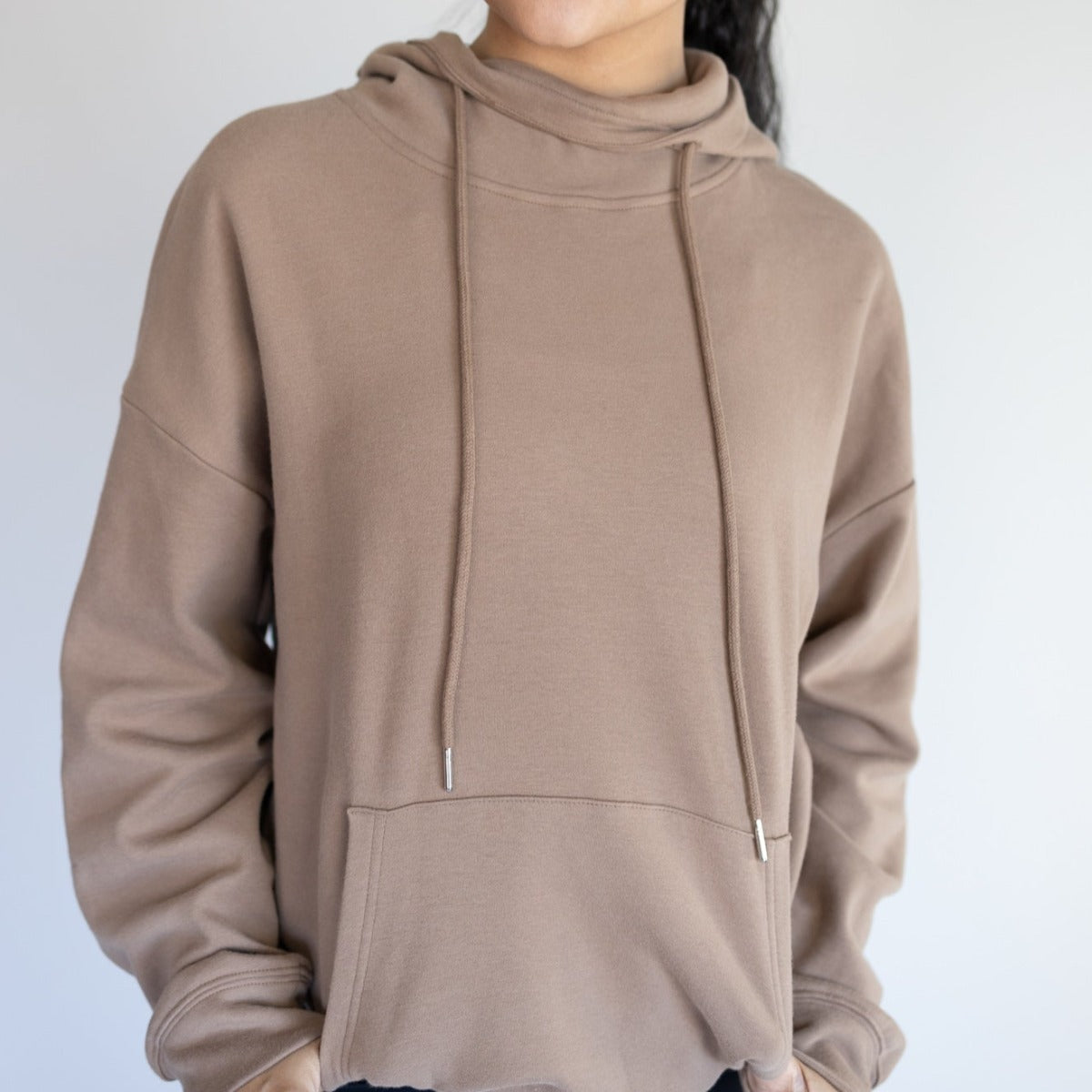 A woman wearing a brown Cozy Up Cowl Neck Hoodie by DYM Athletics, featuring a relaxed fit and stylish cowl design in a bright indoor setting.