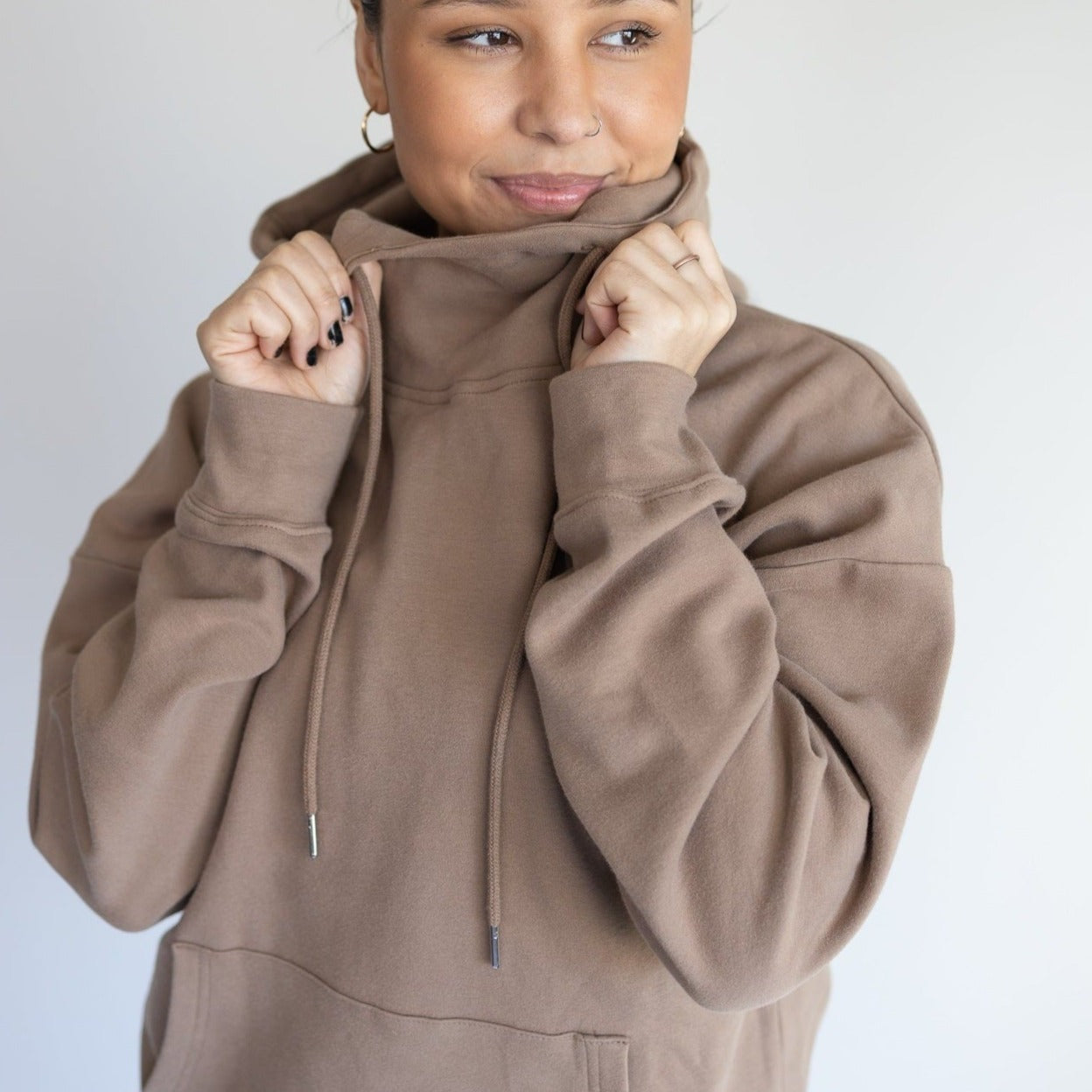 Close-up of a woman playfully holding the collar of her brown Cozy Up Cowl Neck Hoodie from DYM Athletics, emphasizing warmth and comfort.
