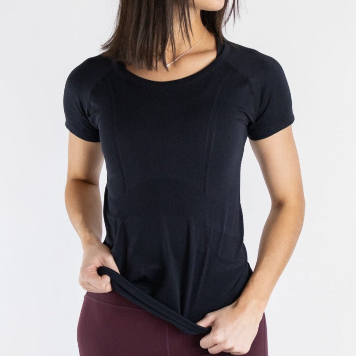 Front view of a woman holding the hem of the Trainer Lightweight Short Sleeve Shirt by DYM Athletics, highlighting its stylish fit and soft fabric.