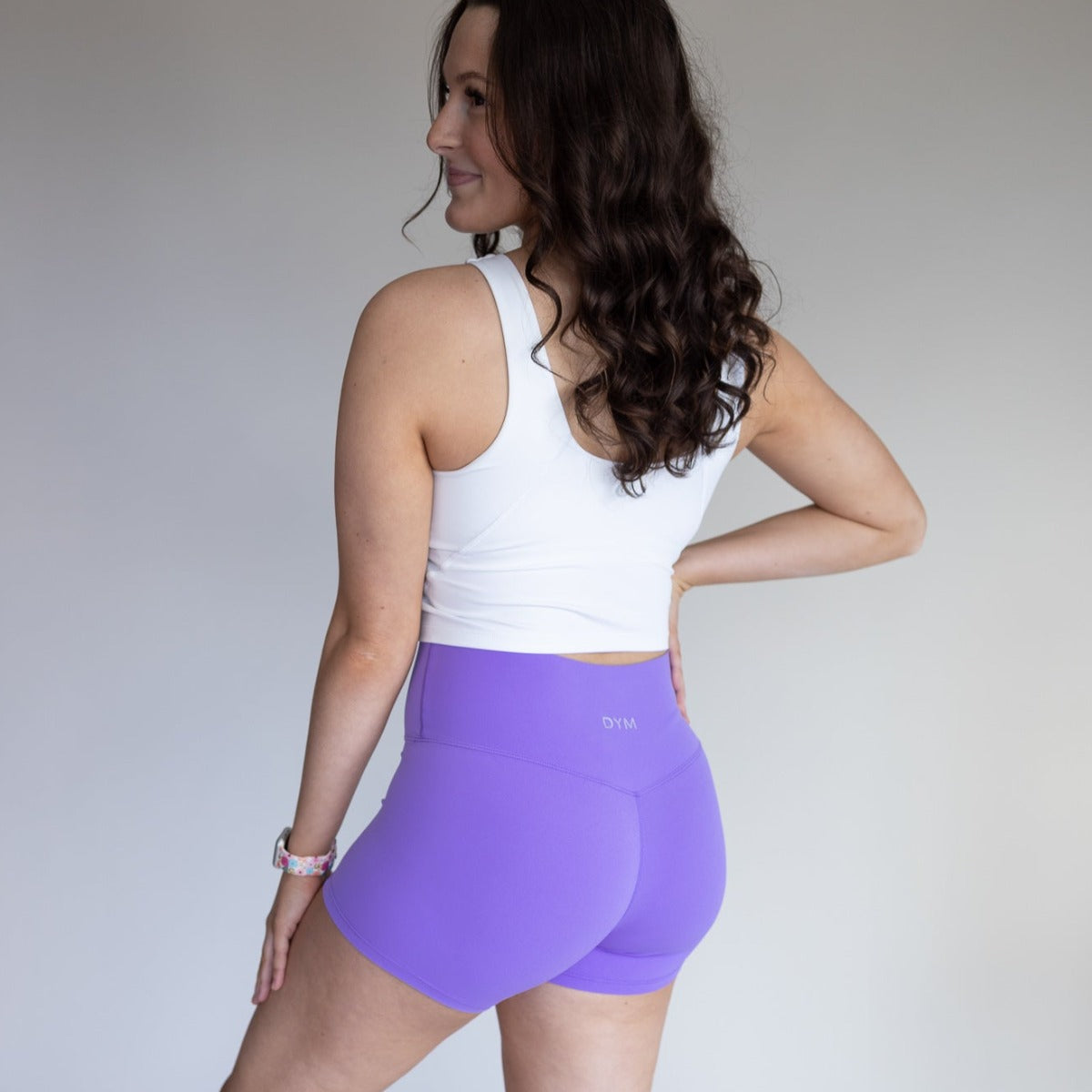 Profile view of a woman wearing a white DYM Athletics Bliss High Neck Bra with Scoop Back, paired with purple shorts, radiating confidence and style.