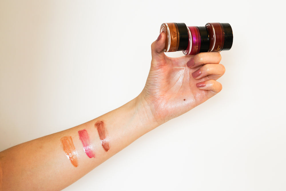 A hand holding True Moringa Lip Whip Trio Set with swatches of the lip whips on the arm, showcasing the vibrant colors and smooth texture.
