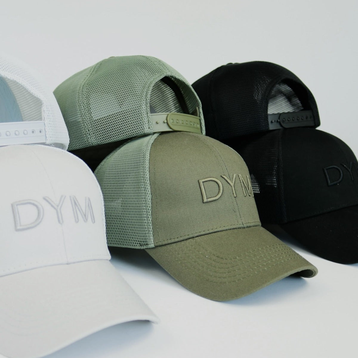 Four DYM Athletics Trucker Hats in different colors including white, olive green, and black stacked together on a white background.