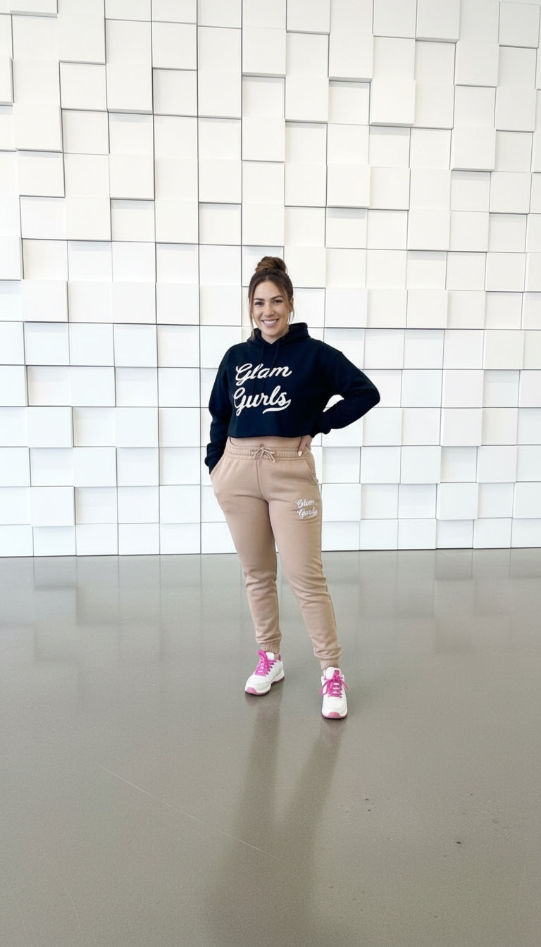 Smiling model confidently posing in a Customized Glam Gurls Cropped Hoodie with Puff Print and beige joggers against a white wall backdrop, highlighting comfort and style. Shop Glam Gurls in sizes S-XL.