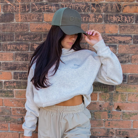 DYM Athletics Trucker Hat showcased on a woman with long hair, styled in a casual outfit against a graffiti wall.