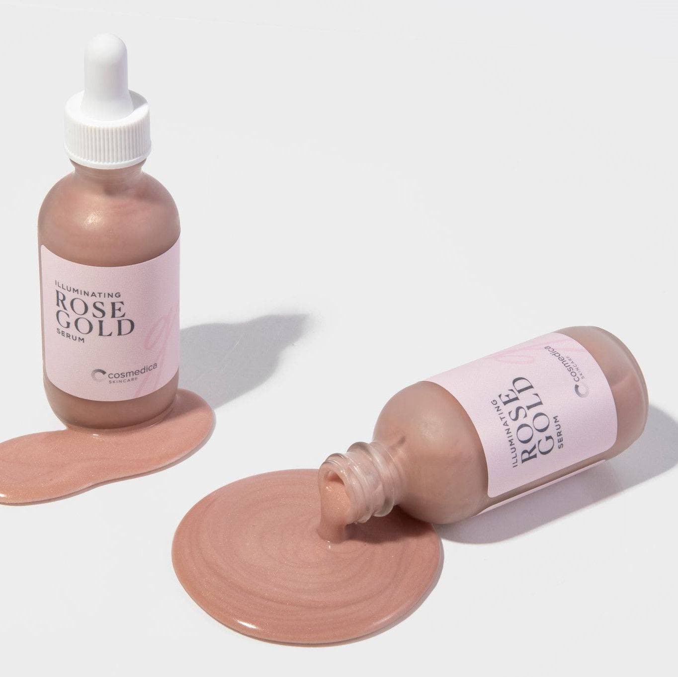 Cosmedica Skincare Illuminating Rose Gold Facial Serum bottles with serum spilled on the surface, showcasing the luxurious texture and signature rose gold hue.