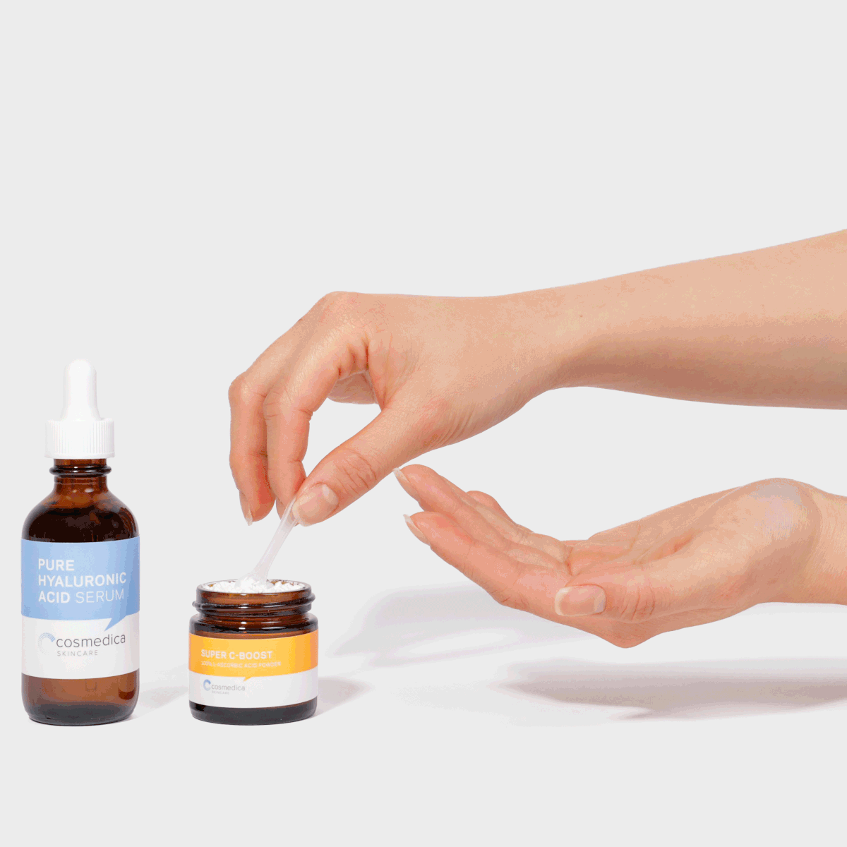 A hand using a spatula to apply Cosmedica Skincare Super C-Boost Vitamin C powder mixed with Pure Hyaluronic Acid serum for a personalized skincare treatment.